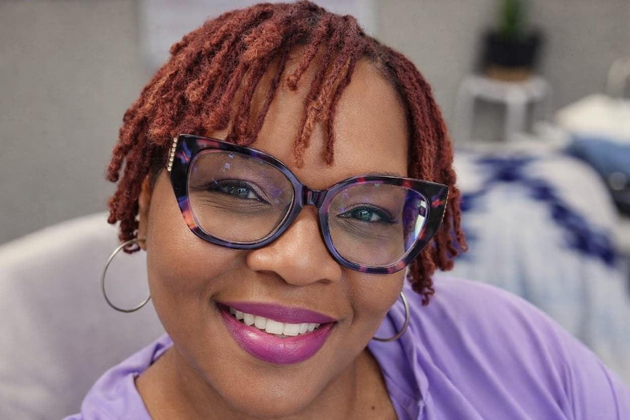 Woman with locs, wearing glasses and a purple shirt with a blurred indoor background