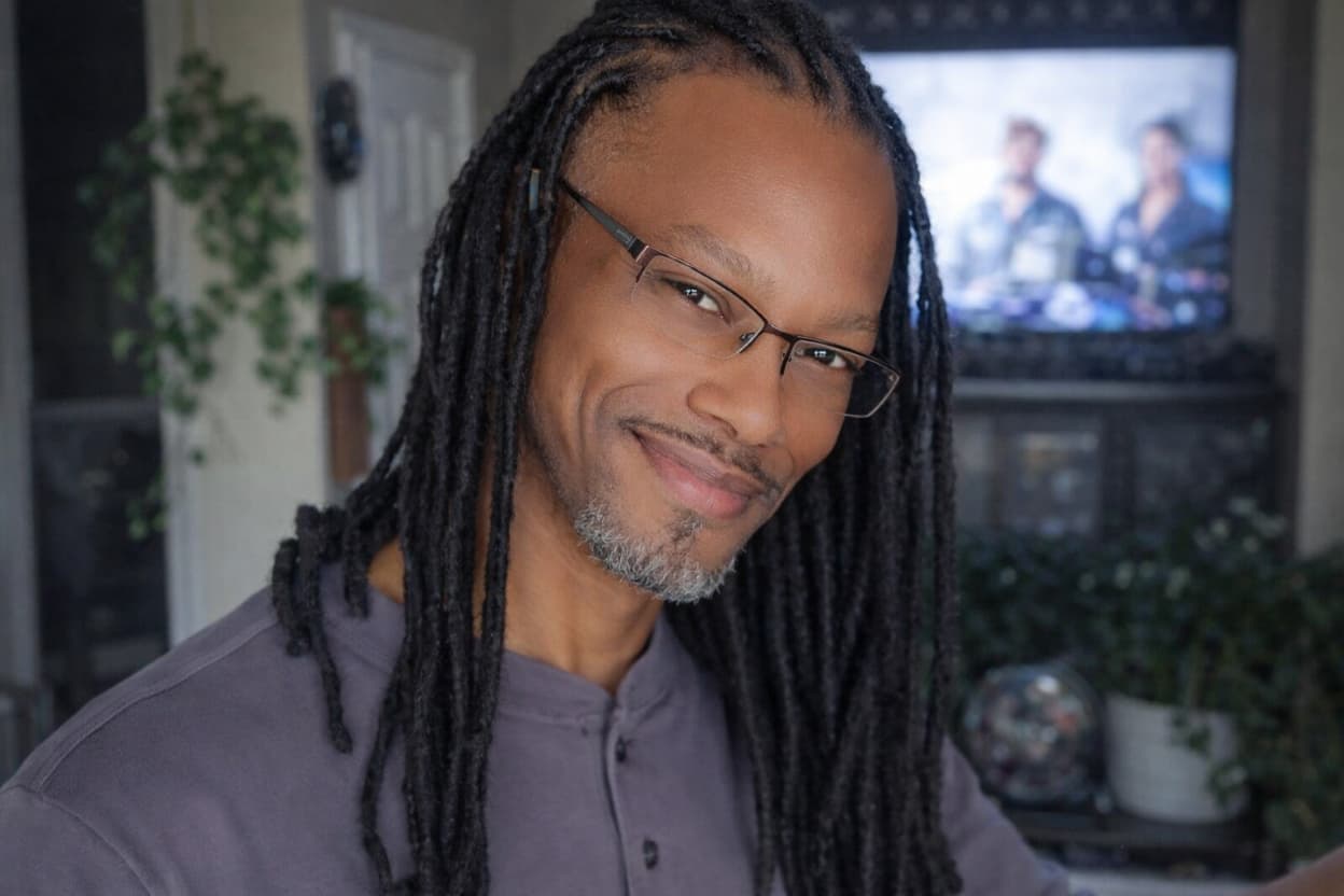 Man with dreadlocks wearing glasses and a purple shirt, sitting in a room with plants and a TV in the background.